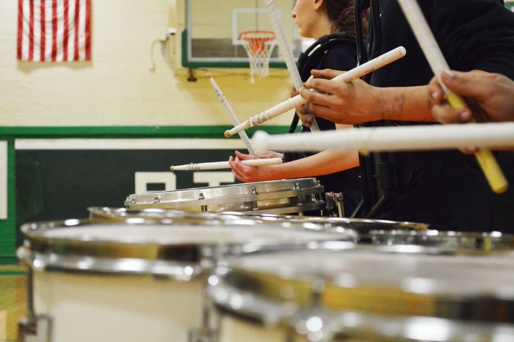 Drumsticks raised over a row of snare drums in a gym