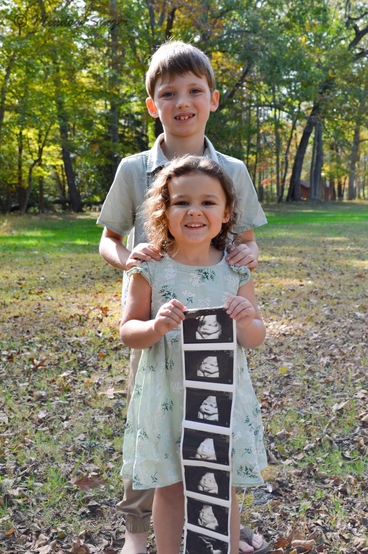 A little girl holding a sonogram strip with her big brother behind her