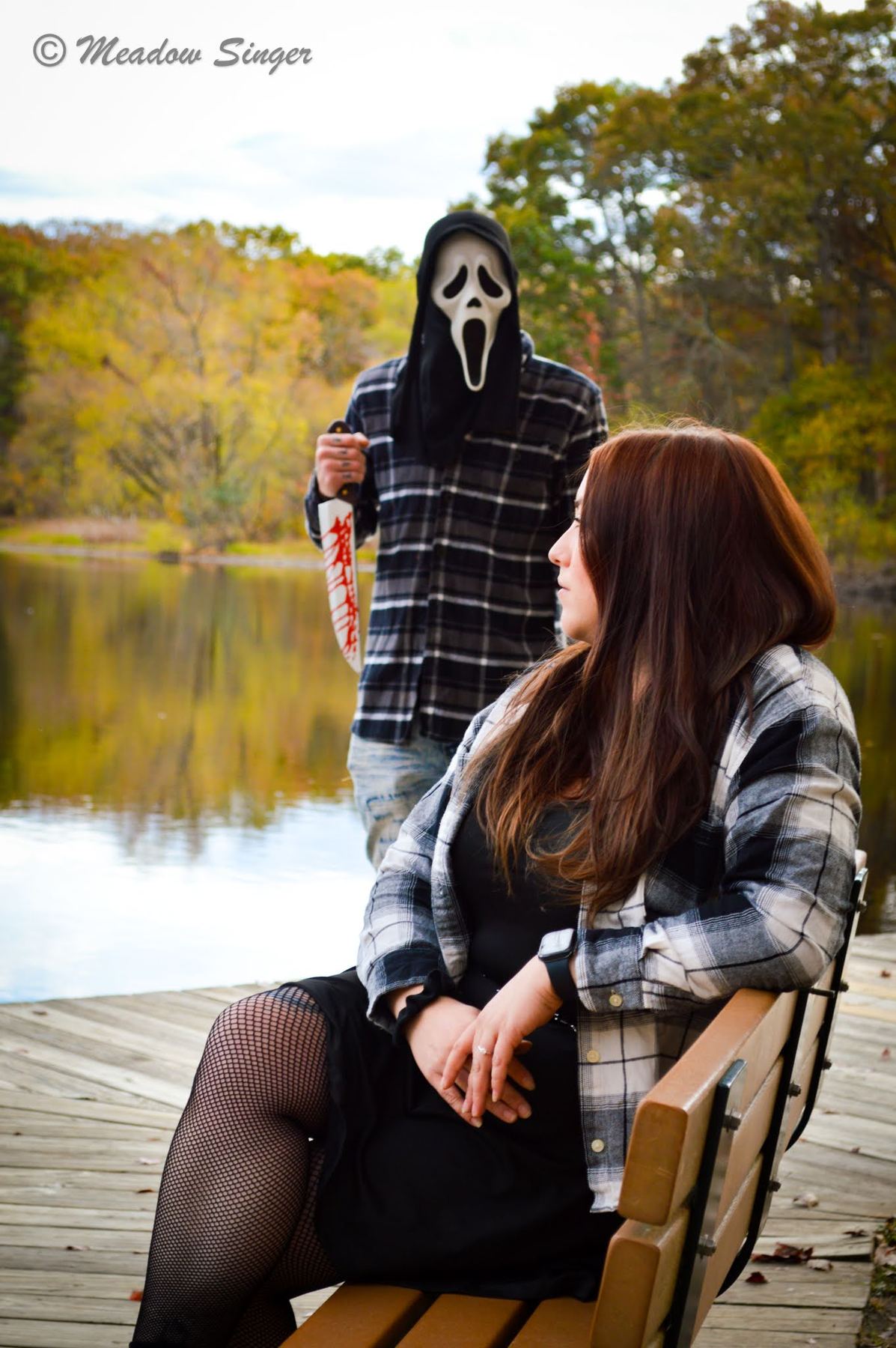 A woman in flannel on a lakeside dock with a masked figure behind her