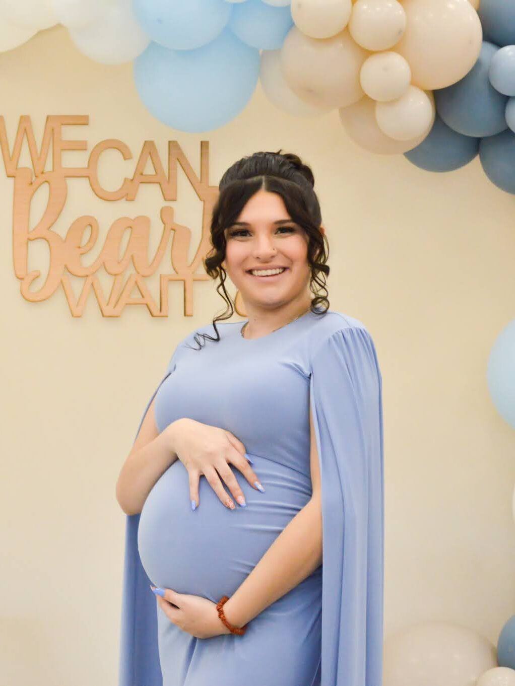An expecting mother smiling in a pale blue gown with a cape